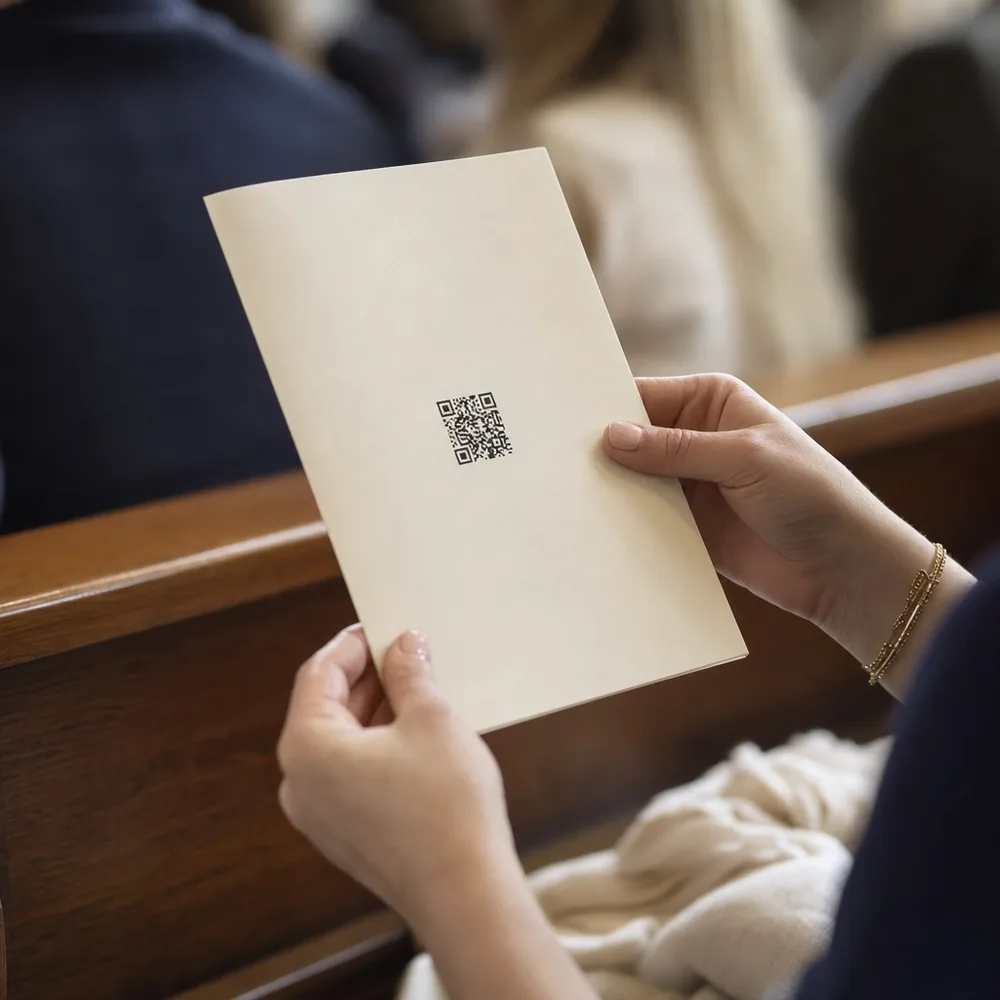 Hands holding a folded ceremony programme turned over to show a small printed QR code on the back, above a wooden pew