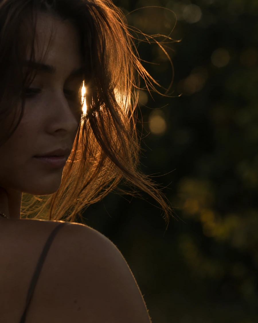 Backlit portrait at golden hour with a thin sliver of sun escaping past the subject's head, throwing warm flare through loose strands of hair