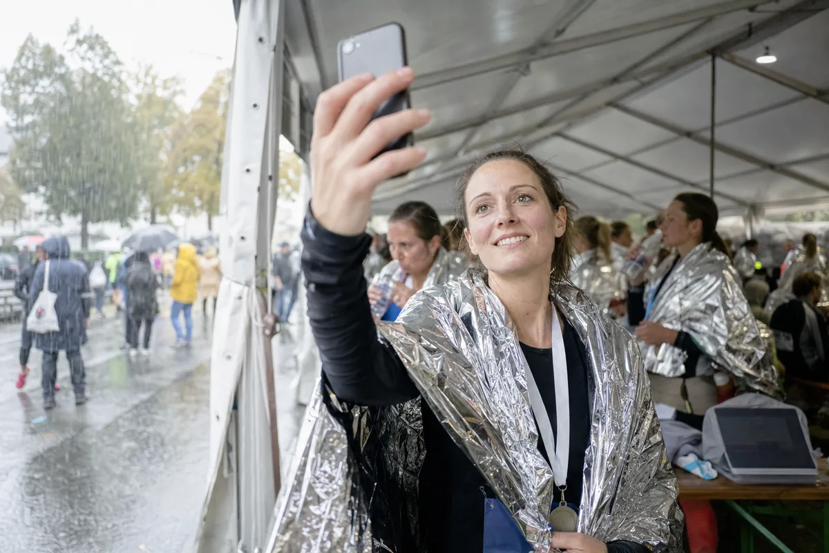 Runner wrapped in a foil blanket under a finish-area tent, holding up her phone to take a selfie while rain falls outside